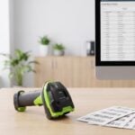 Barcode scanner on a desk beside a monitor showing inventory data and printed barcode labels